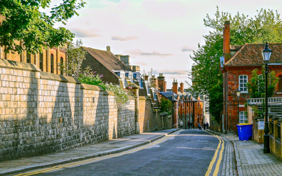 A quiet residential street in Maze Hill with a curved asphalt road marked by double yellow lines. On the left, a tall stone boundary wall runs alongside the pavement, casting shadows from nearby trees