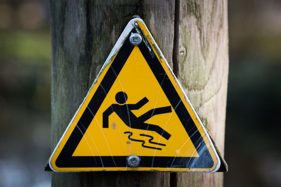 A triangular yellow warning sign with a black border, mounted on a wooden post outdoors in daylight, displays a black pictogram of a person slipping and falling into a puddle, indicating a slippery surface. The sign is attached with metal screws and is slightly weathered, with visible scratches and a faint web of cracks. The background shows out-of-focus natural surroundings with green foliage and possibly water, suggesting an outdoor environment near a path or water hazard. This type of warning sign may be encountered along walkways or outdoor areas, and in the context of home relocations or moving services, it underscores the importance of caution during the loading or transportation process, especially in environments where slippery surfaces could pose risks. Man with Van Maze Hill occasionally incorporates such signage in their safety and logistical planning for house removals and furniture transport, particularly when working in outdoor or variable conditions.