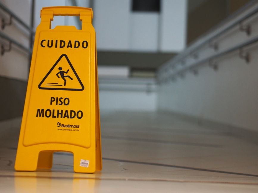 A yellow warning sign with black text and a pictogram, placed on a wooden floor next to a doorway or interior space. The sign reads 'CUIDADO PISO Molhado' (Caution Wet Floor) and features a graphic of a person slipping. It is positioned in a home or building corridor, where the surface appears to be wet, potentially indicating recent cleaning or a hazard. The sign is part of the environment associated with house removals or moving activities, warning occupants or movers of a slippery floor to ensure safety during the packing, loading, or transportation process. The image captures the scene with a focus on safety measures within the context of furniture transport and home relocation, emphasizing the importance of caution in moving environments. Bright lighting highlights the sign and the surrounding area, illustrating typical safety protocols followed during house removals by companies like Man with Van Maze Hill.