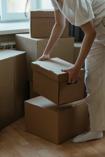 A person wearing light-colored clothing is seen inside a home, arranging and stacking plain brown cardboard boxes on a wooden floor near a window with natural daylight. The individual is holding one box, preparing it for transport during a home relocation process. Several boxes are visible, some labeled with handwritten notes, indicating packed belongings such as clothes. The environment suggests an organized packing scene associated with furniture transport and moving logistics. In the background, a window provides bright natural light, illuminating the packaging area, where the individual is engaged in packing or loading boxes, potentially for a household move managed by Man with Van Maze Hill as part of their removals service. The scene captures the meticulous preparation involved in packing and moving, with the focus on the careful handling of boxes to facilitate a smooth furniture transport and home relocation.