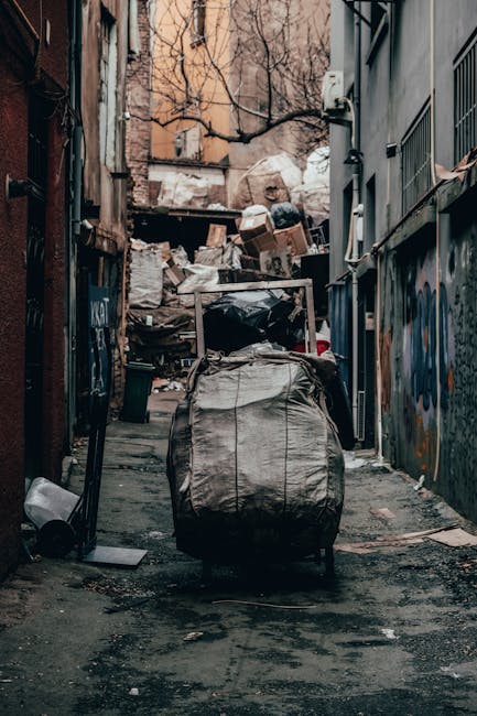 An outdoor alleyway with narrow concrete pavement flanked by tall buildings on both sides, featuring graffiti on the walls and cluttered with various waste and discarded items. In the foreground, a large, weathered fabric bag or sack with visible seams and straps is placed on the ground, possibly containing household waste or debris. Behind it, several cardboard boxes, plastic bags, and other waste materials are piled up against a makeshift fence or wall at the end of the alley. A small plastic waste bin is positioned near the left side of the alley, and some loose trash is scattered around. The scene appears to be part of a waste removal process, potentially related to house moving or clearing a property, with natural lighting casting soft shadows. The presence of a van or vehicle is not visible, but the setting suggests a typical space used for waste or bulky material collection during home relocation. This image reflects the concept of bulky waste removal options in SE10 during house moves, as managed by Man with Van Maze Hill, highlighting the logistical challenges of waste transportation within urban environments.