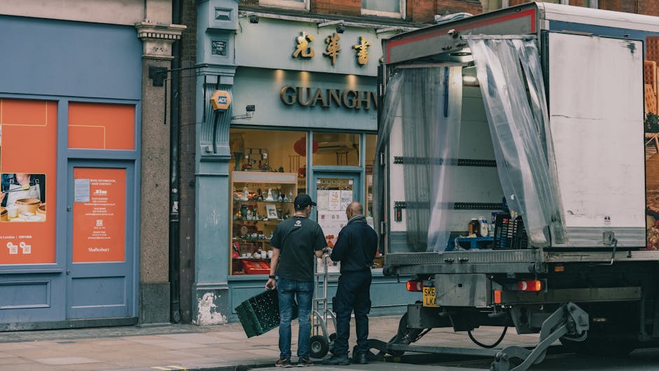 Two movers from Man with Van Maze Hill are loading boxes and small items into the back of a delivery truck during a home relocation process outside a shop in SE10. The front of the truck is open with