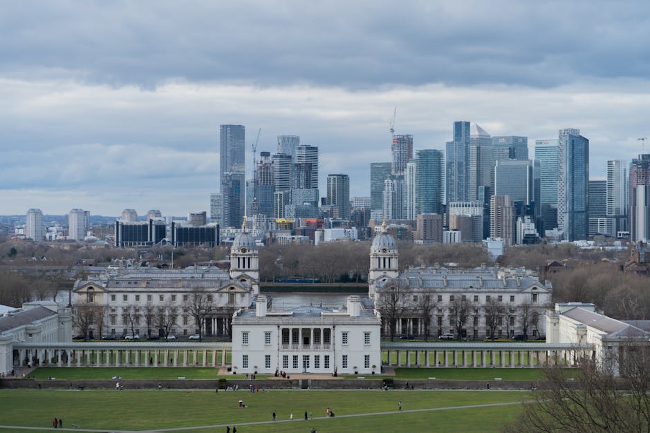 A panoramic view of a historic white mansion with classical architectural features, including multiple wings, columns, and ornate details, set against the backdrop of a modern city skyline with high-rise office buildings under an overcast sky. The mansion is surrounded by a well-maintained lawn with walking paths and a few visitors, and is located within a park area featuring a central green space. The scene captures the contrast between the historic estate and the contemporary urban development in the distance, with visible cranes on some of the skyscrapers indicating ongoing construction. The lighting is natural, highlighting both the detailed facades of the mansion and the sleek, glass surfaces of the modern buildings. This setting illustrates a typical urban landscape where architectural history meets present-day city growth, relevant to house relocations and removals in city environments.
