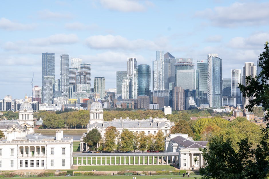 A panoramic view of a historic white mansion with classical architectural features, including multiple wings, columns, and ornate details, set against the backdrop of a modern city skyline with high-rise office buildings under an overcast sky. The mansion is surrounded by a well-maintained lawn with walking paths and a few visitors, and is located within a park area featuring a central green space. The scene captures the contrast between the historic estate and the contemporary urban development in the distance, with visible cranes on some of the skyscrapers indicating ongoing construction. The lighting is natural, highlighting both the detailed facades of the mansion and the sleek, glass surfaces of the modern buildings. This setting illustrates a typical urban landscape where architectural history meets present-day city growth, relevant to house relocations and removals in city environments.