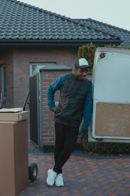 A man wearing a grey cap, black gloves, a dark padded vest over a blue long-sleeve shirt, and black trousers stands outside a residential property, leaning against the open rear door of a white moving