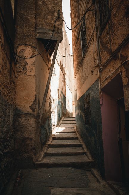 A narrow, cobblestone alleyway between two historic stone buildings, with uneven, aged paving visible on the ground. The walls are constructed from weathered bricks and stones, showing signs of age an