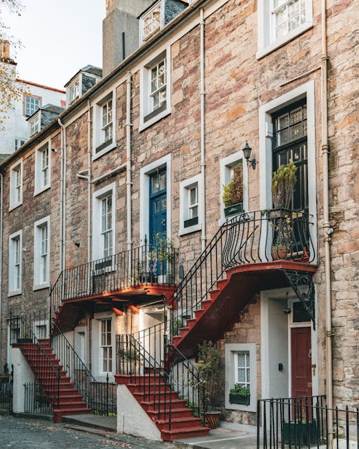The exterior of a historic Georgian townhouse features a stone facade with multiple white-framed sash windows and black wrought-iron railings. A distinctive curved external staircase with red-painted treads and black iron balustrades provides access from the street level to the upper-floor entrance doors, one painted in dark blue and another in black. Potted plants and greenery decorate the staircase landing and window sills, adding a touch of greenery to the building's exterior. The pavement in front of the property is cobbled, and neighboring similar row houses are visible in the background under overcast natural lighting. This scene illustrates the typical architecture of SE10 homes in Maze Hill, highlighting narrow doorways and staircase access often encountered during home relocation and furniture transport processes, as handled by companies like Man with Van Maze Hill, who specialise in removals and moving services in such environments.