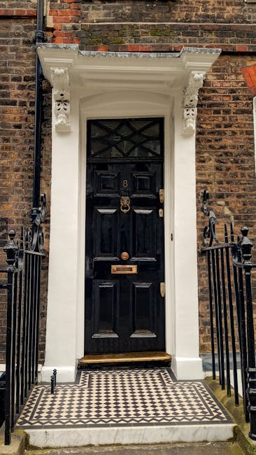 A close-up view of a black wooden front door set within a white classical-style surround on a brick Georgian townhouse in SE10. The door features a brass door knocker, letterbox, and handle, with small glass panels at the top arranged in a diamond pattern. The door is flanked by black wrought-iron railings and entrance steps with a black and white tiled doorstep. There are decorative white brackets supporting the canopy above the door, and the scene is lit with natural daylight. This setting illustrates the challenges of home relocation through narrow doorways typical of Georgian houses, where furniture and packing materials from [COMPANY_NAME] may require careful maneuvering during the loading process for house removals and furniture transport, especially in historic properties with limited access, as highlighted in the 'Narrow doorways in Georgian SE10 homes' page. The image captures the precise environment where professional movers often operate to facilitate efficient packing and moving within constrained entrance spaces.
