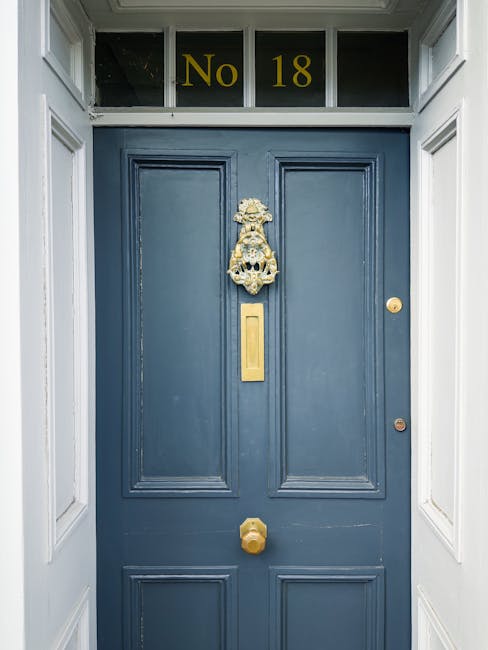 The exterior of a historic Georgian townhouse features a stone facade with multiple white-framed sash windows and black wrought-iron railings. A distinctive curved external staircase with red-painted treads and black iron balustrades provides access from the street level to the upper-floor entrance doors, one painted in dark blue and another in black. Potted plants and greenery decorate the staircase landing and window sills, adding a touch of greenery to the building's exterior. The pavement in front of the property is cobbled, and neighboring similar row houses are visible in the background under overcast natural lighting. This scene illustrates the typical architecture of SE10 homes in Maze Hill, highlighting narrow doorways and staircase access often encountered during home relocation and furniture transport processes, as handled by companies like Man with Van Maze Hill, who specialise in removals and moving services in such environments.