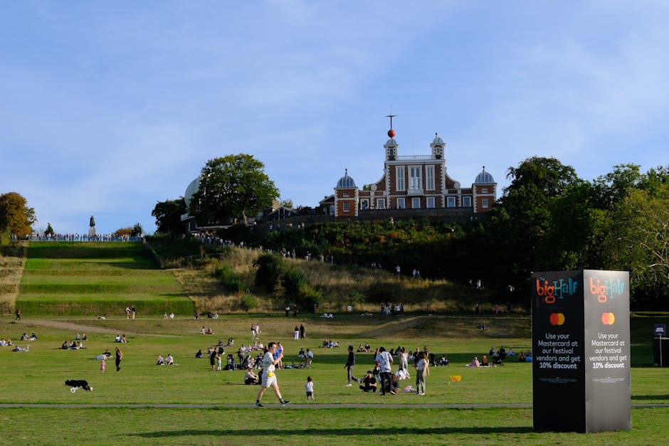 The image depicts a bright, sunny day at a park near the historic Cutty Sark in Greenwich, with a large, open grassy area where numerous people are relaxing, walking, and sitting on the lawn. In the foreground, several individuals and small groups are engaged in leisure activities, some sitting on picnic blankets, others strolling or standing, with a few children and dogs visible. To the right, there's a tall sign advertising a festival with discount offers, and nearby, a variety of cardboard boxes and packing materials are scattered, indicating preparations for home relocation or packing. In the background, a distinctive, ornate building with a symmetrical design and spires, potentially the Cutty Sark museum or a similar historic structure, stands atop a hill surrounded by trees, which cast gentle shadows on the grounds. The scene captures the active process of moving preparations within a lively outdoor environment, with visible equipment like blankets and the scattered packing boxes suggesting ongoing packing and loading activities as part of the moving process.