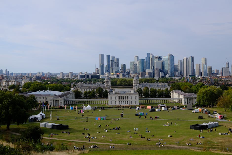 The image depicts a bright, sunny day at a park near the historic Cutty Sark in Greenwich, with a large, open grassy area where numerous people are relaxing, walking, and sitting on the lawn. In the foreground, several individuals and small groups are engaged in leisure activities, some sitting on picnic blankets, others strolling or standing, with a few children and dogs visible. To the right, there's a tall sign advertising a festival with discount offers, and nearby, a variety of cardboard boxes and packing materials are scattered, indicating preparations for home relocation or packing. In the background, a distinctive, ornate building with a symmetrical design and spires, potentially the Cutty Sark museum or a similar historic structure, stands atop a hill surrounded by trees, which cast gentle shadows on the grounds. The scene captures the active process of moving preparations within a lively outdoor environment, with visible equipment like blankets and the scattered packing boxes suggesting ongoing packing and loading activities as part of the moving process.