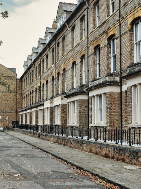 A row of three terraced houses with brick and painted facades, featuring bay windows on the ground and upper floors. The houses are situated on a paved street with a cyclist parked in front near a tra